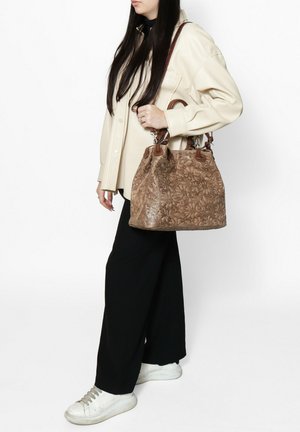 Tan floral-embossed leather handbag with brown accents, dual handles, and a structured shape, worn over a cream shirt and black pants.