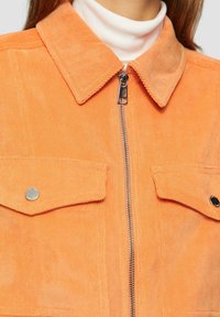 Oranje corduroy jasje met een puntkraag, ritsluiting en twee borstzakken met zilveren knopen. Textiel met een gestructureerd oppervlak en verticale ribbels.