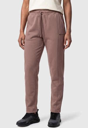 Personne portant un pantalon de jogging mauve à cordon, des baskets noires et un haut blanc, debout devant un fond clair uni.