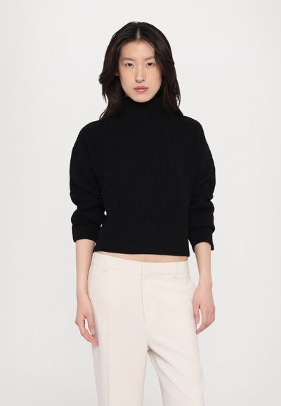 Black ribbed turtleneck sweater with cropped length and voluminous sleeves, paired with light beige trousers.