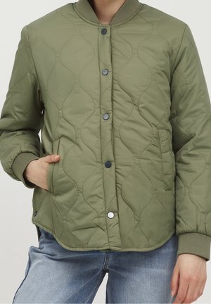 Veste matelassée vert olive avec couture ondulée, fermeture à boutons-pression, col et poignets côtelés, portée avec un jean bleu. Une main dans la poche.