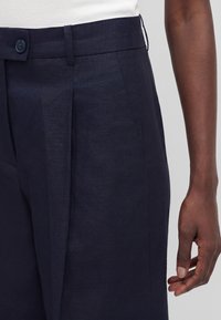 Pantalon bleu marine foncé ajusté avec des plis à l'avant et une fermeture par bouton, porté avec un haut blanc et un bras droit visible détendu.