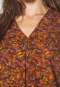 Vestido de tela marrón con estampado, con escote en V y diseños florales en amarillo, azul y verde, con una superficie ligeramente texturizada.