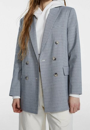 Dobbeltbrystet blazer i blå og hvid houndstooth-mønster, med sølvknapper, lapels med hak og sidelommer, lagdelt over en hvid hoodie.