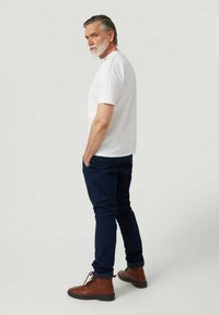 Camiseta blanca de algodón, pantalones azul marino, botas de cuero marrón. El modelo está de lado con las manos en los bolsillos, mangas cortas, textura suave.