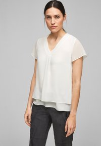Blusa bianca, leggera, a maniche corte con scollatura a V e silhouette stratificata, caratterizzata da morbide pieghe nella zona del collo. Tessuto liscio.