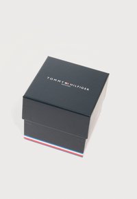 Σκούρο μπλε κουτί ρολογιού Tommy Hilfiger με το λογότυπο στην κορυφή και κόκκινες, λευκές και μπλε ρίγες στη βάση, πάνω σε λευκό φόντο.