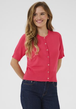 Cardigan corail ajusté à manches courtes, avec un design à boutons sur le devant, texture côtelée et tissu lisse. Porté par-dessus un jean noir.