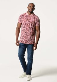 Korte mouwen polo in het roze met een bloemmotief, gecombineerd met blauwe jeans en witte sneakers. Heeft een kraag en ribbels aan de randen.