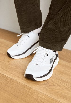 Białe buty sportowe z siateczkową cholewką, czarnymi akcentami i teksturowaną podeszwą. Sznurowadła są związane. Noszone z zielonymi spodniami z kordurowej tkaniny na drewnianej podłodze.