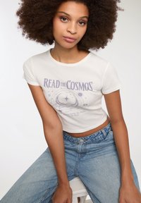 Biała, krótka koszulka z purpurowym napisem "CZYTAJ KOSMOS" oraz motywami niebieskimi, zestawiona z wysokotaliowymi dżinsami w kolorze niebieskim.