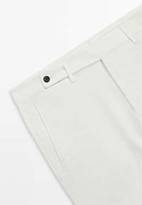 Białe spodnie z płaskim przodem, dwiema kieszeniami po bokach i pojedynczym czarnym guzikiem w talii. Gładka tekstura i dopasowany krój.