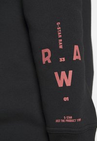 Černá mikina s růžovým textem "G-Star RAW," "33," "01," a "Just the Product 1989." Má měkkou texturu a žebrované manžety.