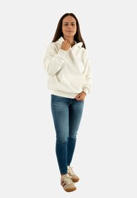 Femme debout portant un hoodie blanc, un jean bleu et des baskets beiges, tenant le col du hoodie d'une main et l'autre à hauteur de taille.