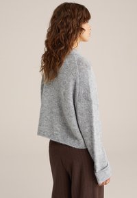 Sivkasta skrajšana jopica, izdelana iz mehkega, puhastega materiala, z rebrastimi detajli na rokavih in ramenih. Ima sproščen kroj in minimalen dizajn.