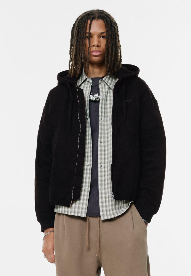 Sort zip-up hættetrøje lagd ovenpå en lys grøn og hvid ternet skjorte, parret med en mørkegrå T-shirt og beige bukser.