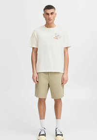 Cremefarvet bomulds t-shirt med rund hals og korte ærmer, med et farverigt grafisk design på venstre bryst. Sættes sammen med beige shorts.