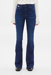 Σκούρο μπλε τζιν καμπάνα με ψηλή μέση. Κατασκευασμένο από υφάσμα denim, διαθέτει μπροστινές τσέπες και κλείσιμο με κουμπί. Λεία υφή.