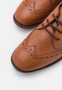 Hnědé kožené dress boty typu wingtip s dekorativními perforacemi a tmavě hnědými tkaničkami na bílém pozadí.