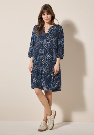 Blau gemustertes Kleid mit V-Ausschnitt, Dreiviertelärmeln und einem gestuften Rock. Kombiniert mit beigen Sneakers für einen lässigen Look.