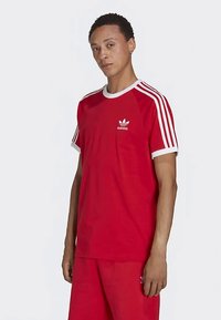 Rdeča bombažna majica z belimi poudarki, okrogel izrez in trije beli trakovi na vsakem ramenu. Na prsih je prikazan Adidasov logotip.
