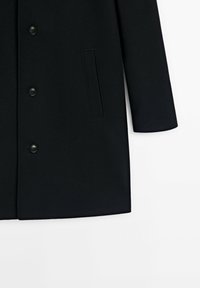 Manteau noir sur mesure avec trois boutons noirs, une manche longue visible et une poche fendue verticale sur le côté droit.