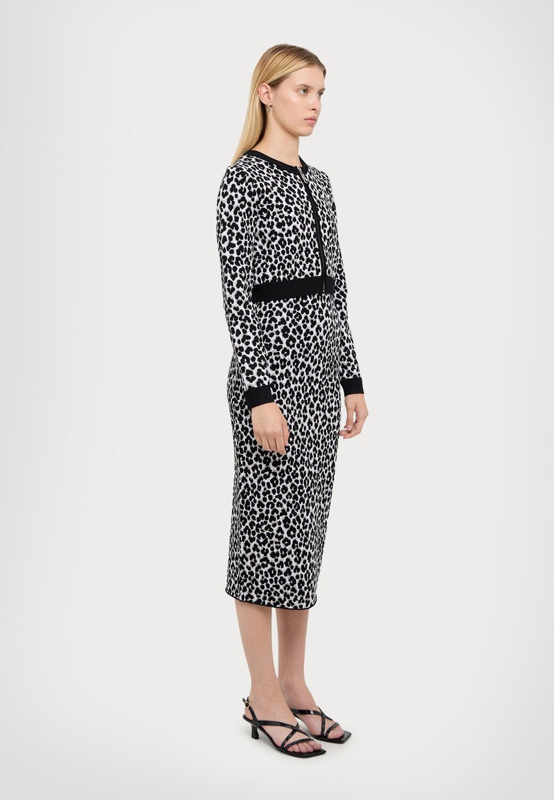 Robe à imprimé léopard en noir et blanc, design ajusté avec une taille noire accentuée et des manches longues. Associée à des sandales noires à lanières.