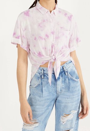 Frau trägt ein kurzärmeliges, hellrosa Batik-Button-up-Hemd, das an der Taille gebunden ist, mit zerrissenen, hellblauen, hoch taillierten Jeans.