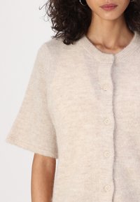 Cardigan beige a maniche corte realizzato in un materiale morbido e peloso, con un collo rotondo e cinque bottoni abbinati lungo il davanti.