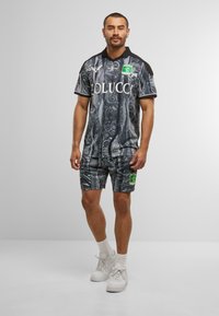Mann steht in einem schwarz-grau gemusterten Sporttrikot und passenden Shorts mit grün-gelbem Logo, weißen Socken und weißen Turnschuhen.