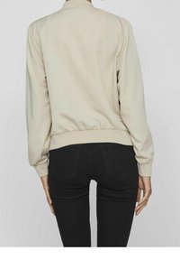 Beige bomberjakke med ribbet krave og cuffs, elastisk talje; sat sammen med sorte tætsiddende jeans, vist fra ryggen. Silket tekstur.
