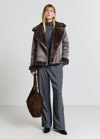 Veste en shearling grise avec un large col, associée à un pull gris texturé et un pantalon large à motifs. Tenant un sac en suède marron.