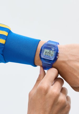 Digitalna ura s prozornim modrim plastičnim ohišjem in trakcem. Kvadratna številčnica prikazuje čas in datum, ter ima srebrne poudarke in logotip.