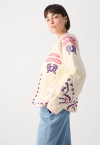 Quiltet kremgenser med rosa og lilla blomsterdesign; har rund halslinning, lange ermene og diamantmønster tekstur.