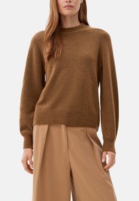 Femme portant un pull en tricot à manches longues marron et un pantalon large beige, se tenant avec les bras détendus sur les côtés devant un fond clair.