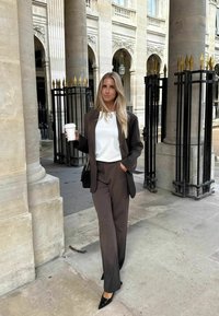Hnědý oblek na míru s bílou košilí, černé lodičky s špičatou špičkou a černá kabelka. Stojící v architektonickém prostředí s sloupy.