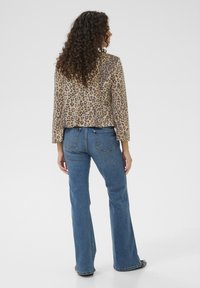Beige Jacke im Leopardenprint mit schwarzen Flecken; kombiniert mit blauen Schlagjeans, von hinten präsentiert, um Textur und Schnitt zu betonen.