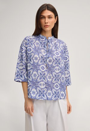 Frau mit glattem braunem Haar trägt eine blau-weiß gemusterte Bluse mit Blumenmotiv und Dreiviertelärmeln sowie weiße Hose, steht vor einfachem Hintergrund.