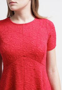 Röd klänning med korta ärmar som har ett strukturerat blommönster, rundad halsringning och figursydd överdel med utsvingad nederdel.