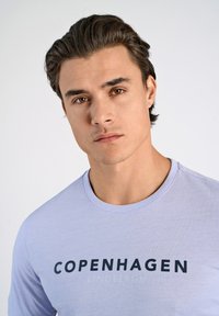 Vaalea violetti pitkähihaine paita, jossa on pyöreä kaula-aukko ja rohkeaa tummansiniistä tekstiä "COPENHAGEN LINDBERGH" rinnan kohdalla. Sujuva puuvillakangas.