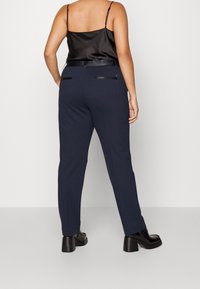 Navy Hose aus Stretchstoff, taillierter Schnitt, zwei Gesäßtaschen mit schwarzen Akzenten, kombiniert mit einem schwarzen Satin-Camisole und klobigen Absätzen.