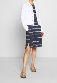 Vit jeansjacka över en marinblå tie-dye klänning med ränder. Klänningen har en sidoflik. Beige sandaler fulländar looken.