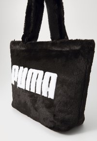Sac fourré noir en simili cuir avec deux poignées robustes, comportant un grand logo "PUMA" blanc sur un côté. Texture douce et forme rectangulaire.