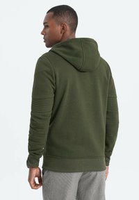 Sudadera verde oliva con acabado texturizado, detalles acanalados en las mangas y un diseño ajustado. Cuenta con un bolsillo tipo canguro en la parte frontal y capucha ajustable.