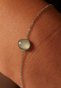 Zilveren kettingarmband met een doorzichtig groene ovale steen, gezet in decoratief metalen omhulsel. De armband ligt op een gestructureerde huidoppervlak.