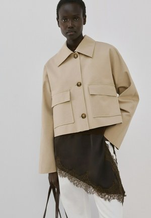 Mujer de piel oscura que lleva una chaqueta corta beige con grandes bolsillos, una falda oscura con encaje y sostiene un bolso marrón oscuro.