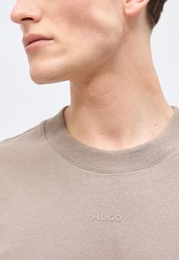 Κοντινό πλάνο της γνάθου και του λαιμού ενός ατόμου που φοράει μπεζ μπλούζα με στρογγυλή λαιμόκοψη και το λογότυπο "HUGO" κεντημένο στο στήθος.