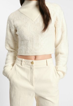 Žena v krémovém krátkém pleteném svetru s texturovaným vzorem a vysokým pasem krémových manšestrových kalhot s knoflíkovým zapínáním, ruce v kapse.