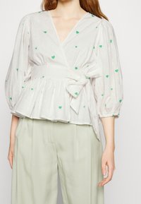 Vit blus med gröna hjärtan, korta puffärmar, bältesmidja och peplumform, kombinerad med ljusgröna byxor.