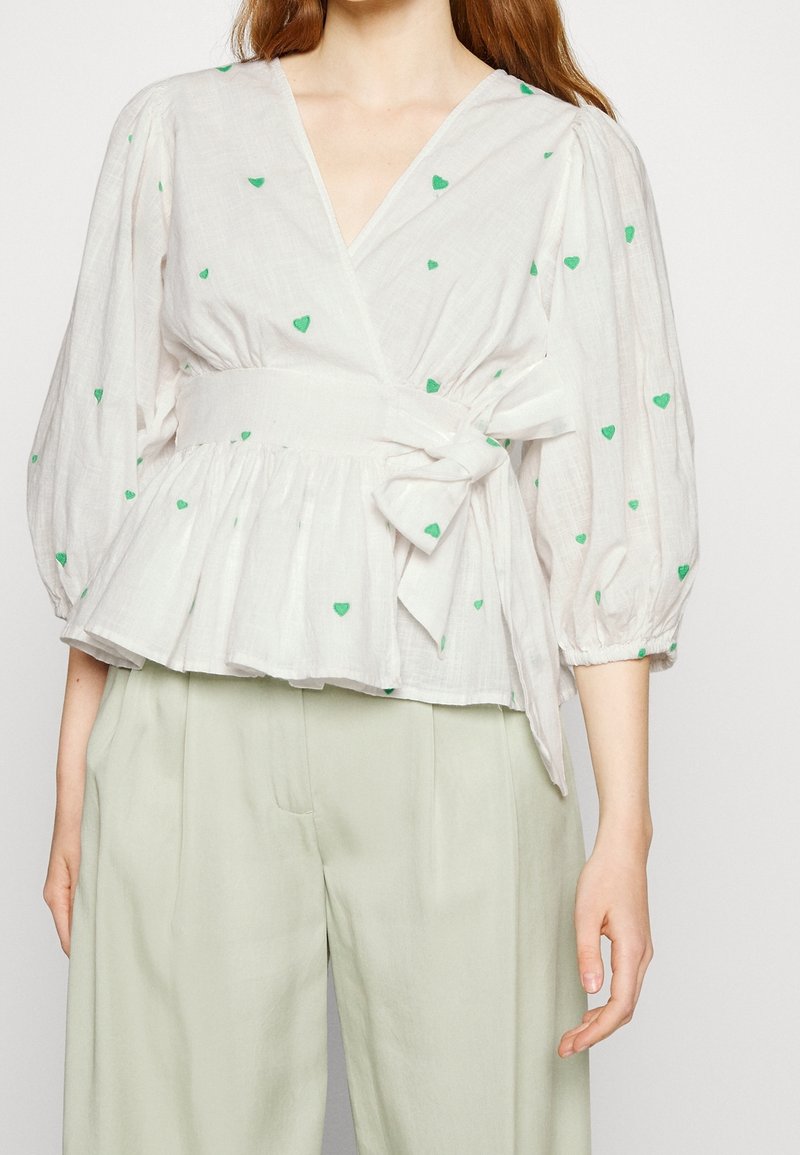 Vit blus med gröna hjärtan, korta puffärmar, bältesmidja och peplumform, kombinerad med ljusgröna byxor.